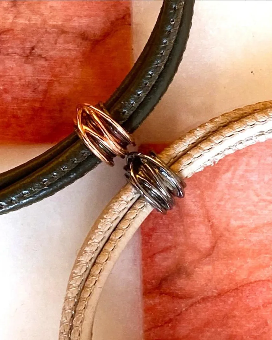 Dettaglio di due bracciali in pelle, uno chiaro e uno scuro, con passanti a spirale in oro rosa e argento brunito